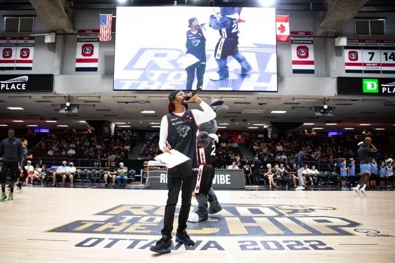 A person drinking from a bottle while standing on a basketball court at the 2022 OTTAWA CHAMPS event. A mascot and a large screen displaying live footage are visible in the background, along with seated spectators and banners.