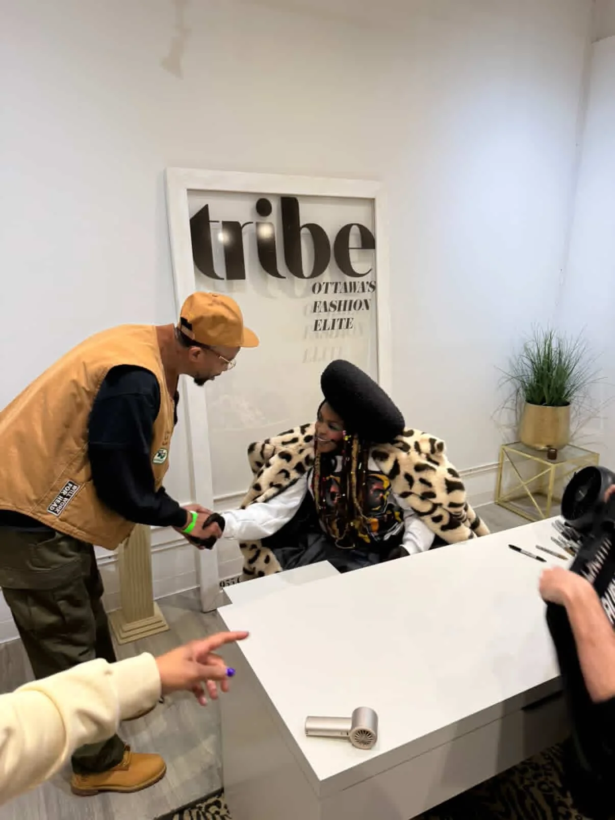 A woman with a large black hairdo, wearing a leopard-print coat, sitting at a white desk and shaking hands with a man wearing a tan cap and vest. There are two other people's hands visible, one pointing at the woman and the other's holding a camera. 