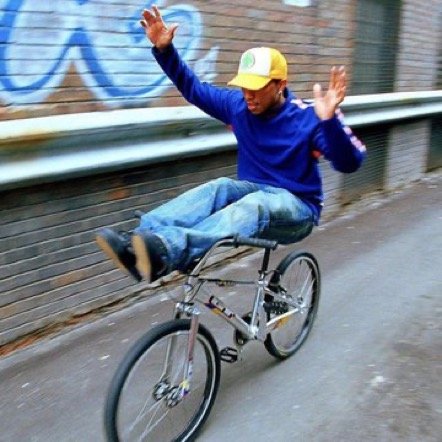 Person riding a bicycle with their arms raised, wearing a yellow cap, blue jacket, and jeans, on a city sidewalk.