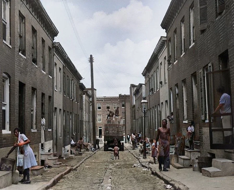 A neighborhood under construction with brick row houses and people working and walking along the unpaved street.