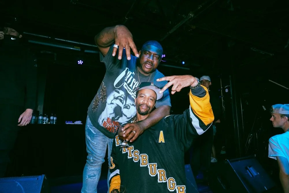 Two men posing together on a stage, one standing and one sitting. The man standing has his hand on the other's head, making a peace sign, both smiling. The man sitting is wearing a baseball jersey with "PITTSBURGH" on it and a cap. The background sho