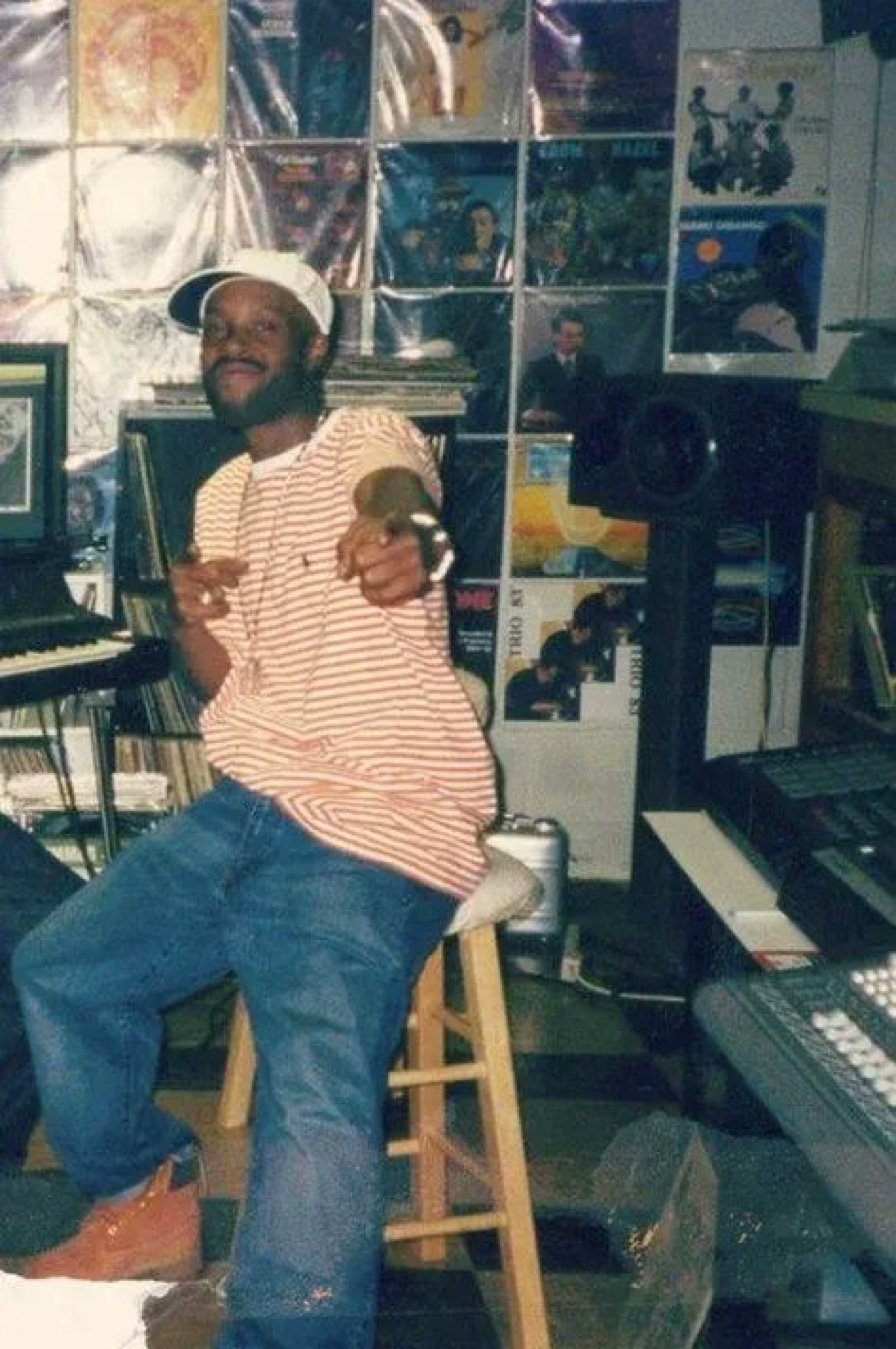 A young man sitting on a wooden stool in a music studio, pointing towards the camera with a smile. He is wearing a striped t-shirt, jeans, a white cap, and brown boots. The studio has musical equipment, vinyl records, posters, and a wall made of mirr
