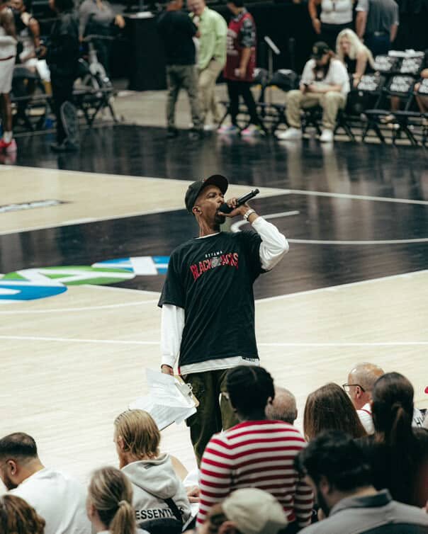 A man in a black cap and black t-shirt with red text on the back, standing on a basketball court, speaking or singing into a microphone, during a public event.
