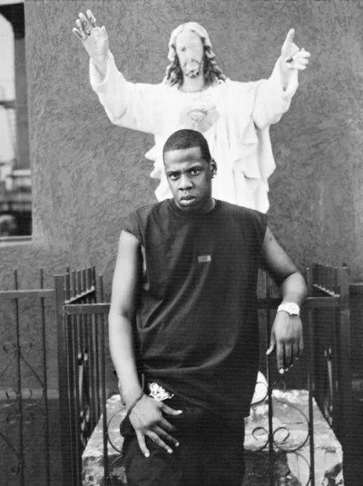 A young man posing in front of a statue of Jesus with arms raised and a peaceful expression.