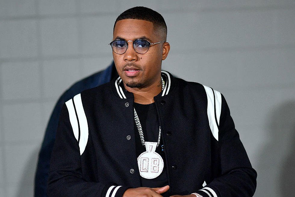 A man with short hair, wearing blue glasses, a black and white varsity jacket, and a large pendant necklace.