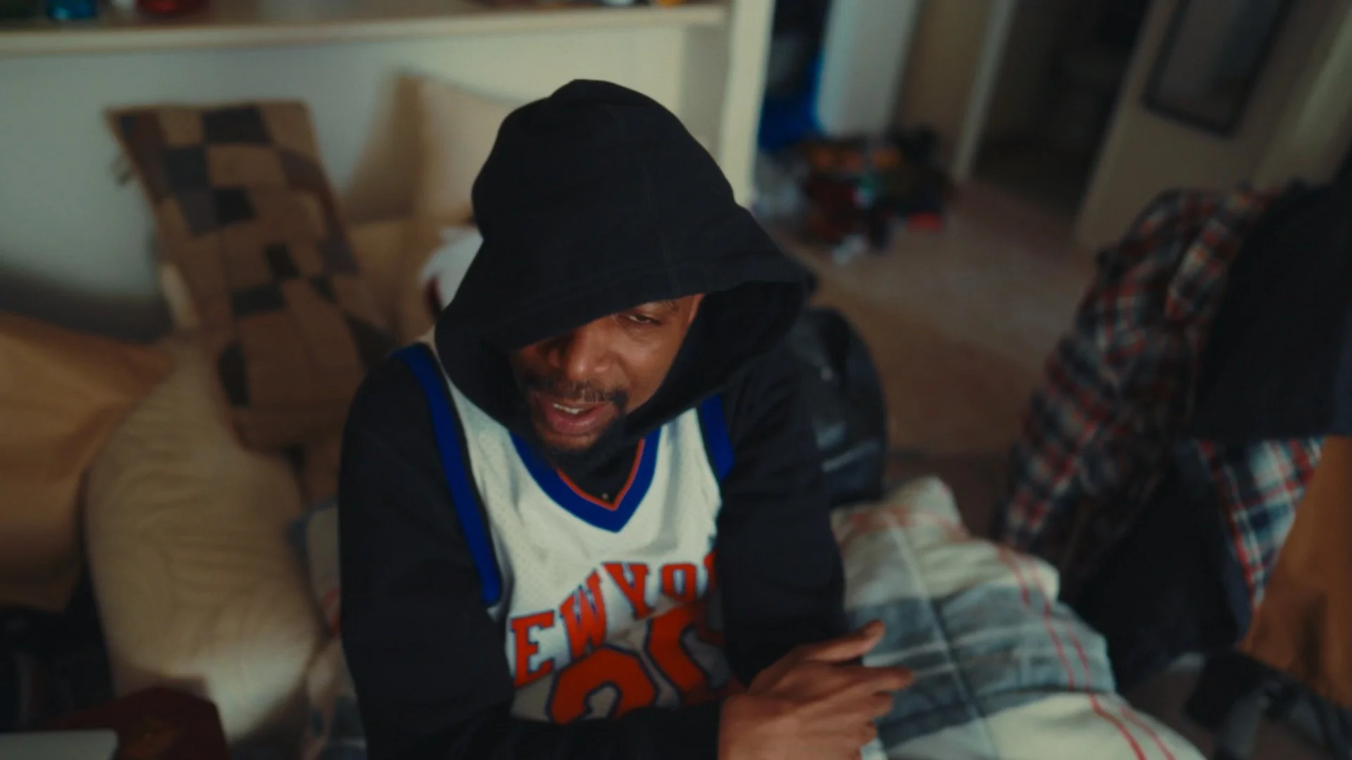 A man wearing a hoodie and a basketball jersey sitting inside a room, appearing distressed or emotional.