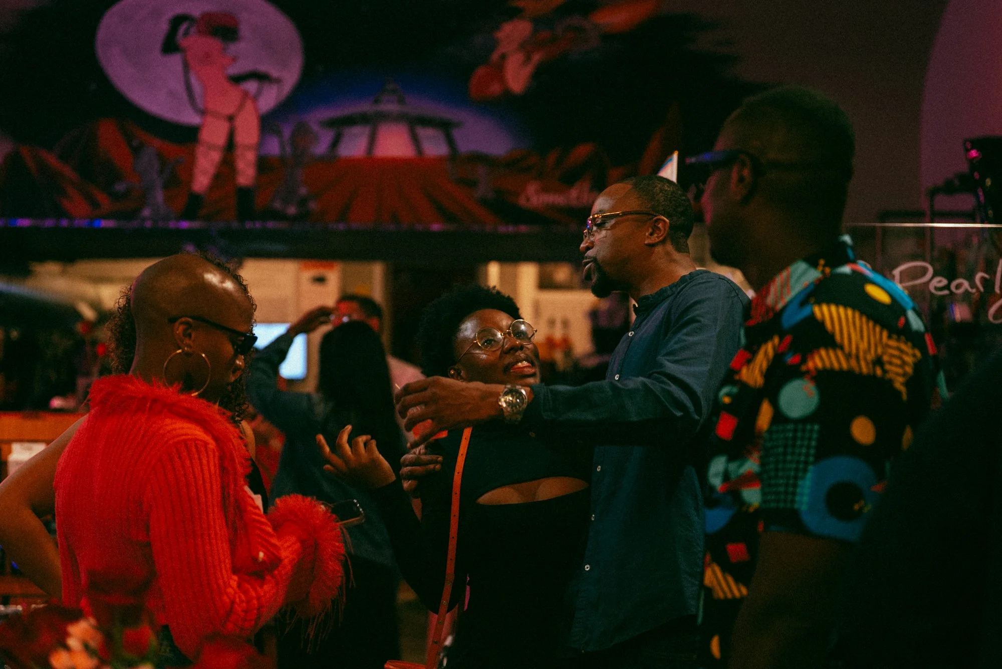People engaging in conversation at a dimly lit indoor event with vibrant, colorful decor in the background.