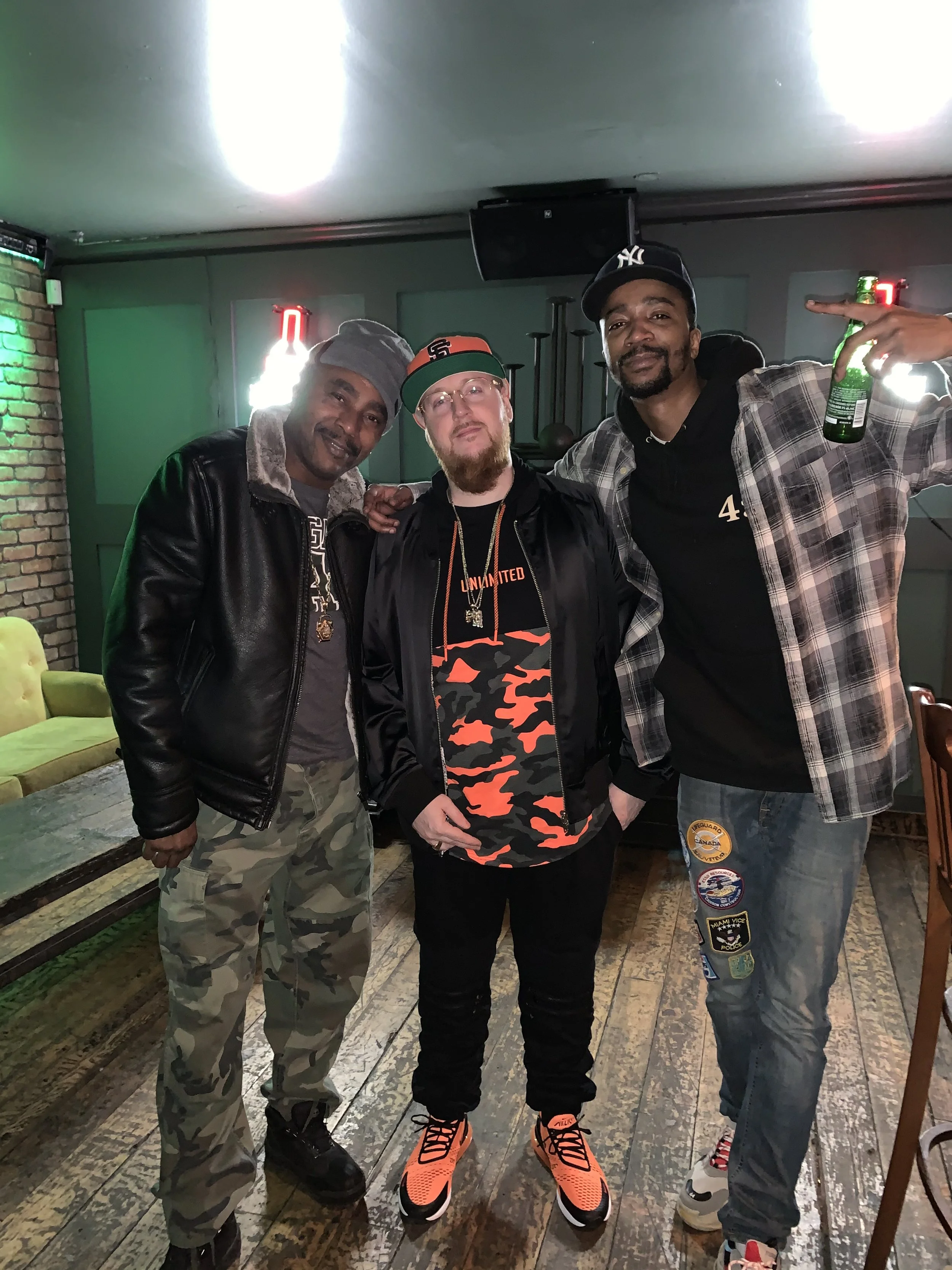 Three men standing together in a bar, smiling at the camera. The man on the left is wearing a leather jacket, camouflage pants, and a gray beanie. The man in the middle has a beard, wearing a black jacket, red and black camo t-shirt, and orange sneak