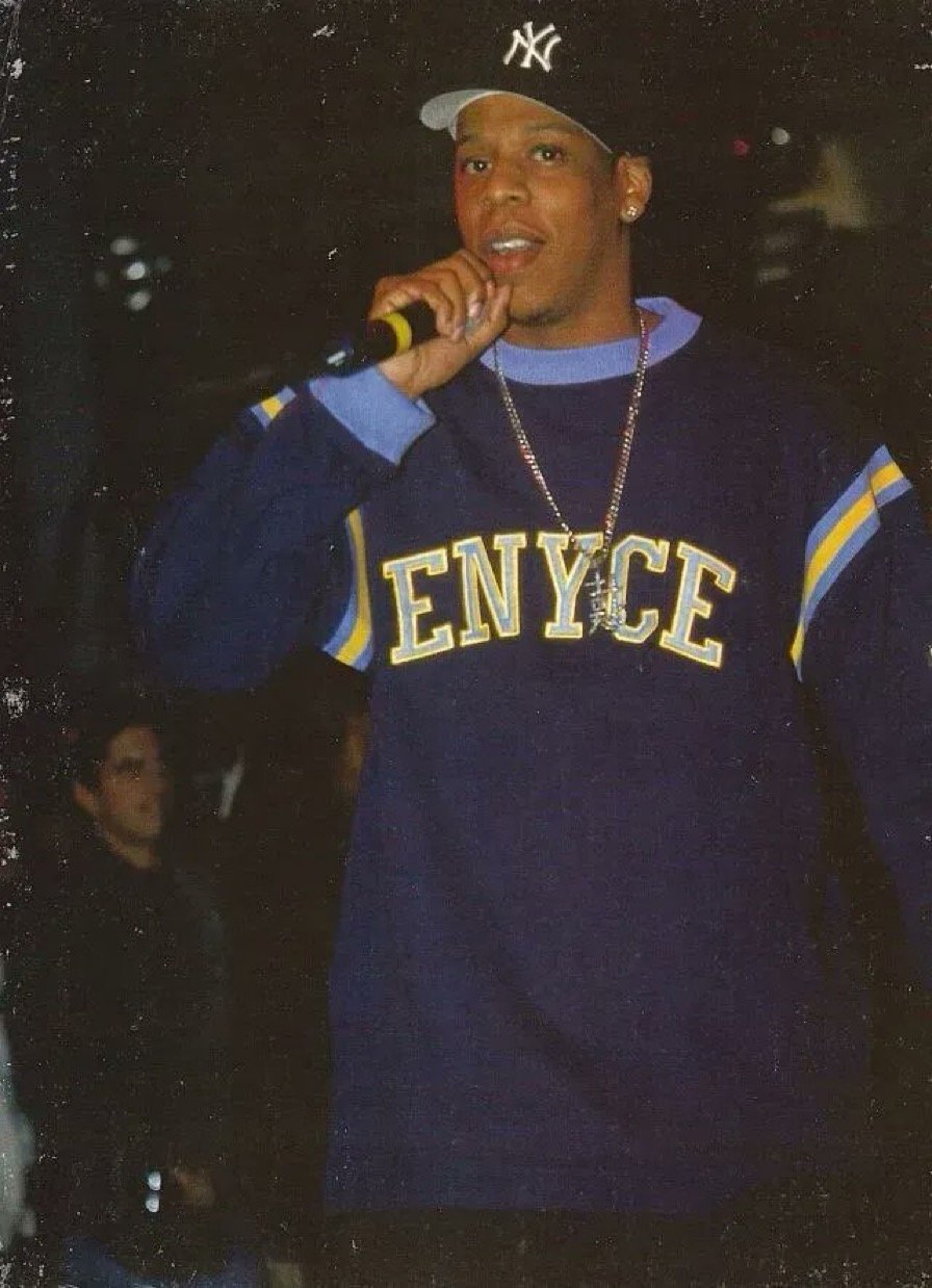 Young man in a blue NY Yankees cap and navy blue sweatshirt with 'ENYCE' logo, holding a microphone, performing on stage.