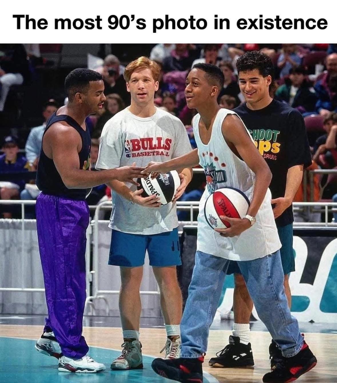 Four young men on a basketball court, talking and holding basketballs, with a crowd of spectators in the background.