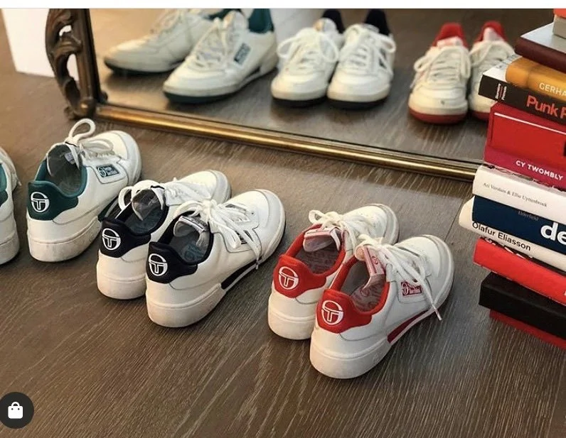 Several pairs of white sneakers with different colored accents (teal, black, red) arranged on a wooden surface in front of a mirror, with a stack of books to the right.