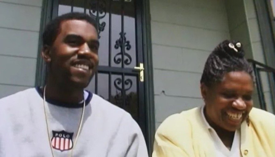 Two people sitting outdoors near a house with a decorative metal security door. The man on the left is smiling, wearing a light gray sweatshirt with a logo and a necklace. The woman on the right is laughing, wearing a yellow cardigan and having her h