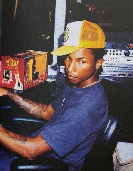 A young man with tattoos wearing a yellow trucker hat and a blue T-shirt, sitting in a recording studio surrounded by audio equipment and a box with 'The Muppet Show' branding.