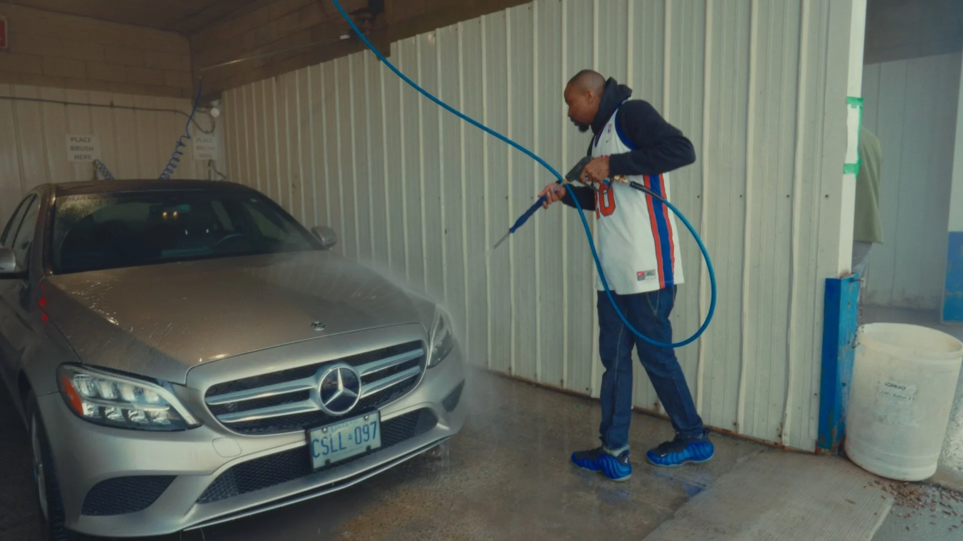 Man washing a gray Mercedes-Benz car with a hose at a car wash station.