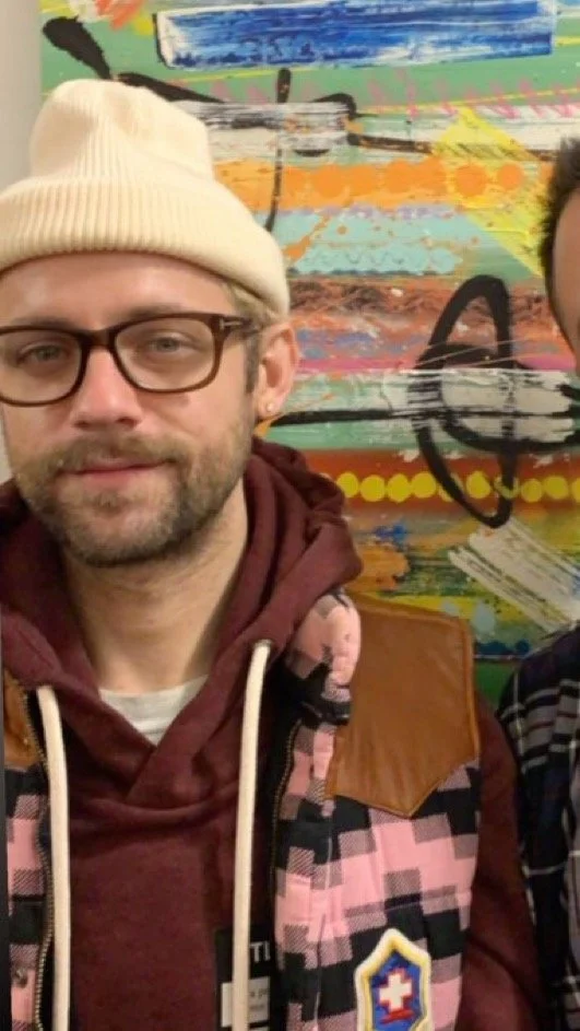 A man wearing a beige beanie, glasses, a maroon hoodie, and a patterned vest stands in front of a colorful, abstract mural.