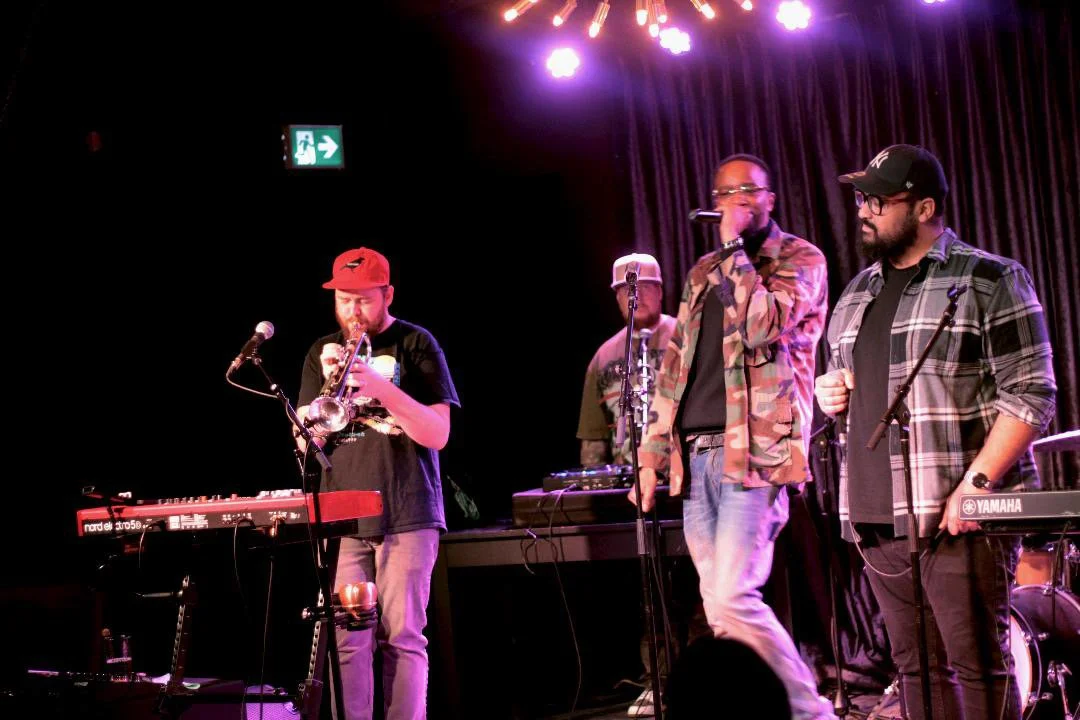 A music band performing on stage with three members singing into microphones and playing instruments, including a keyboard and a trumpet, under stage lights with a dark curtain background.