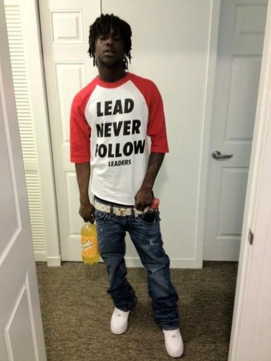 Young man with dreadlocks wearing a red and white baseball shirt that says "LEAD NEVER FOLLOW LEADERS," holding a yellow drink in his left hand, standing in a doorway in a home, with a red item in his right pocket and white sneakers.