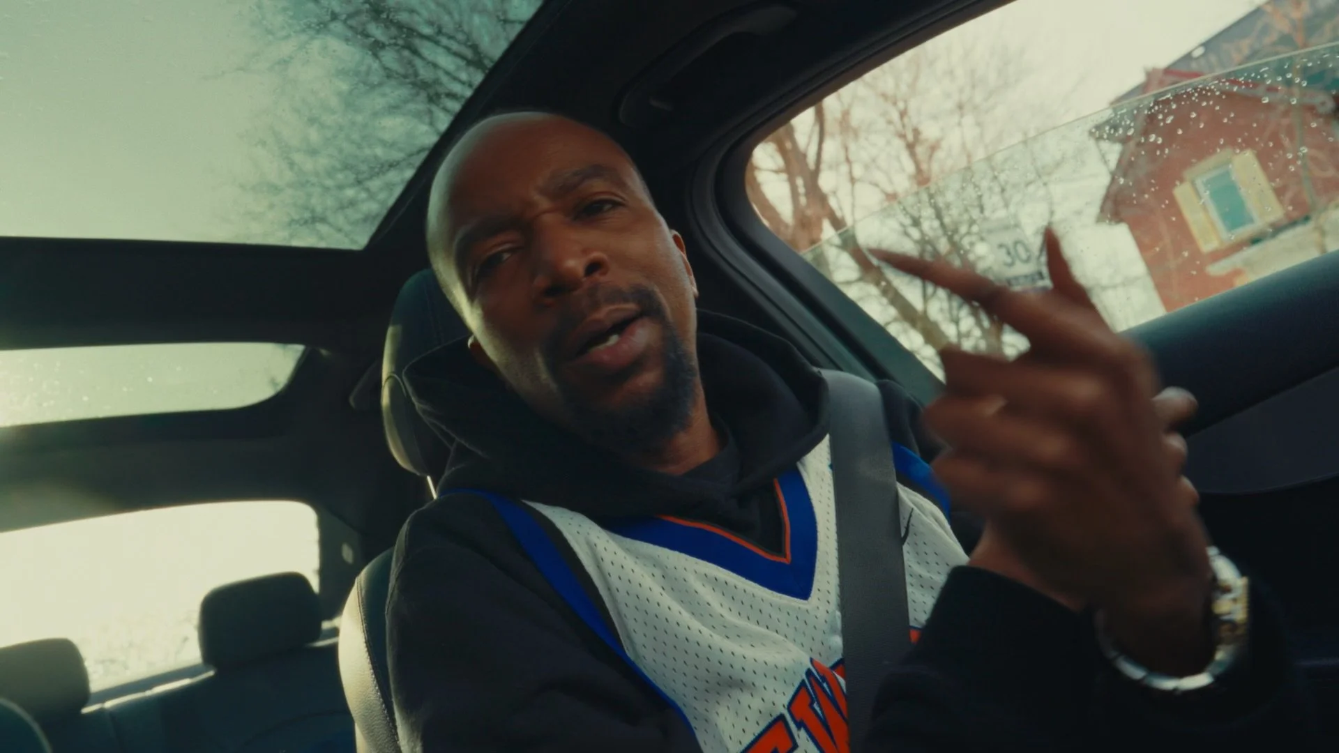 A man with a shaved head and goatee wearing a sports jersey inside a car, talking and gesturing with his hand, with trees and buildings visible through the windows.