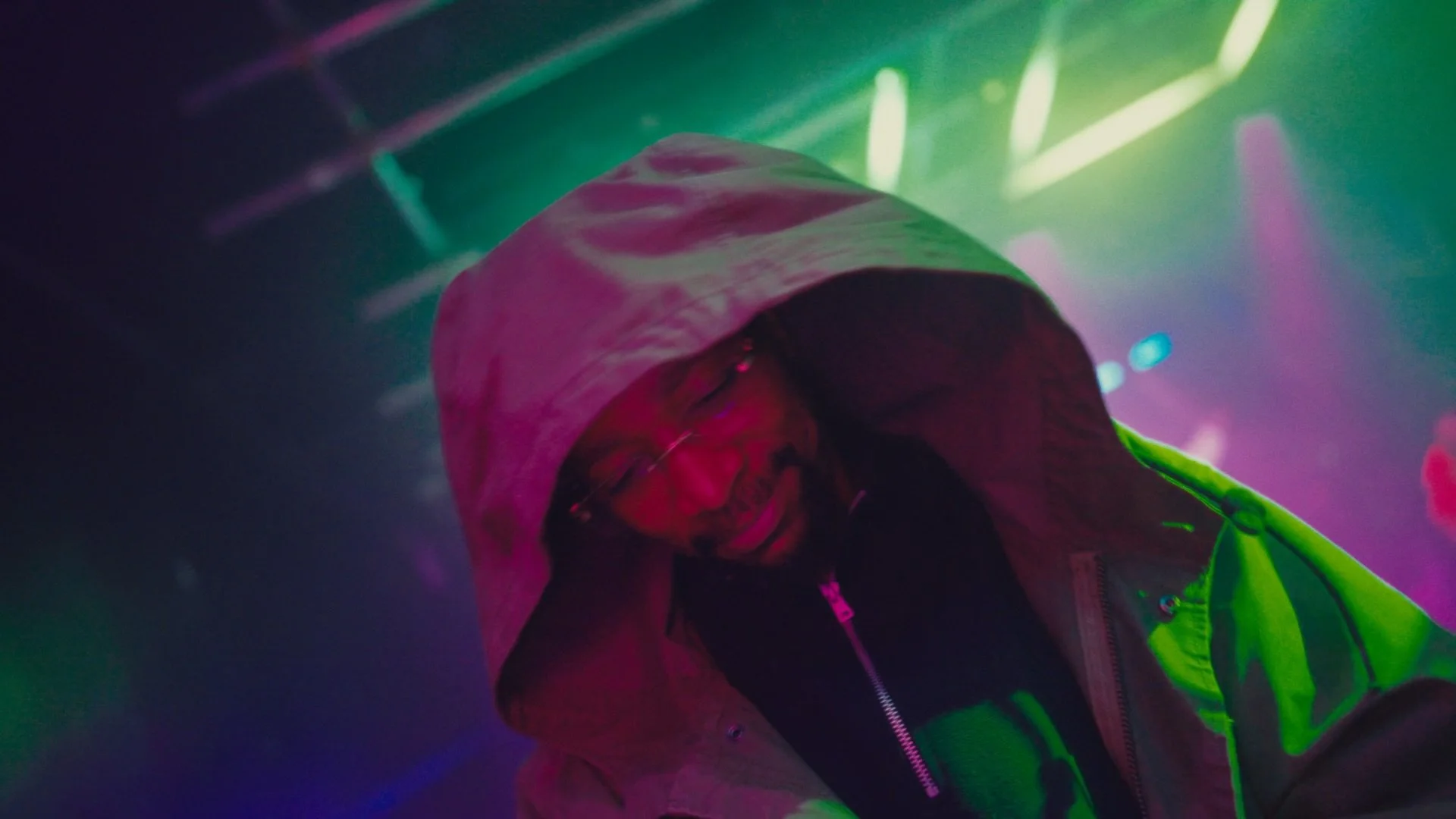 Person wearing glasses and a hooded jacket, smiling, surrounded by colorful lights in a dark setting.