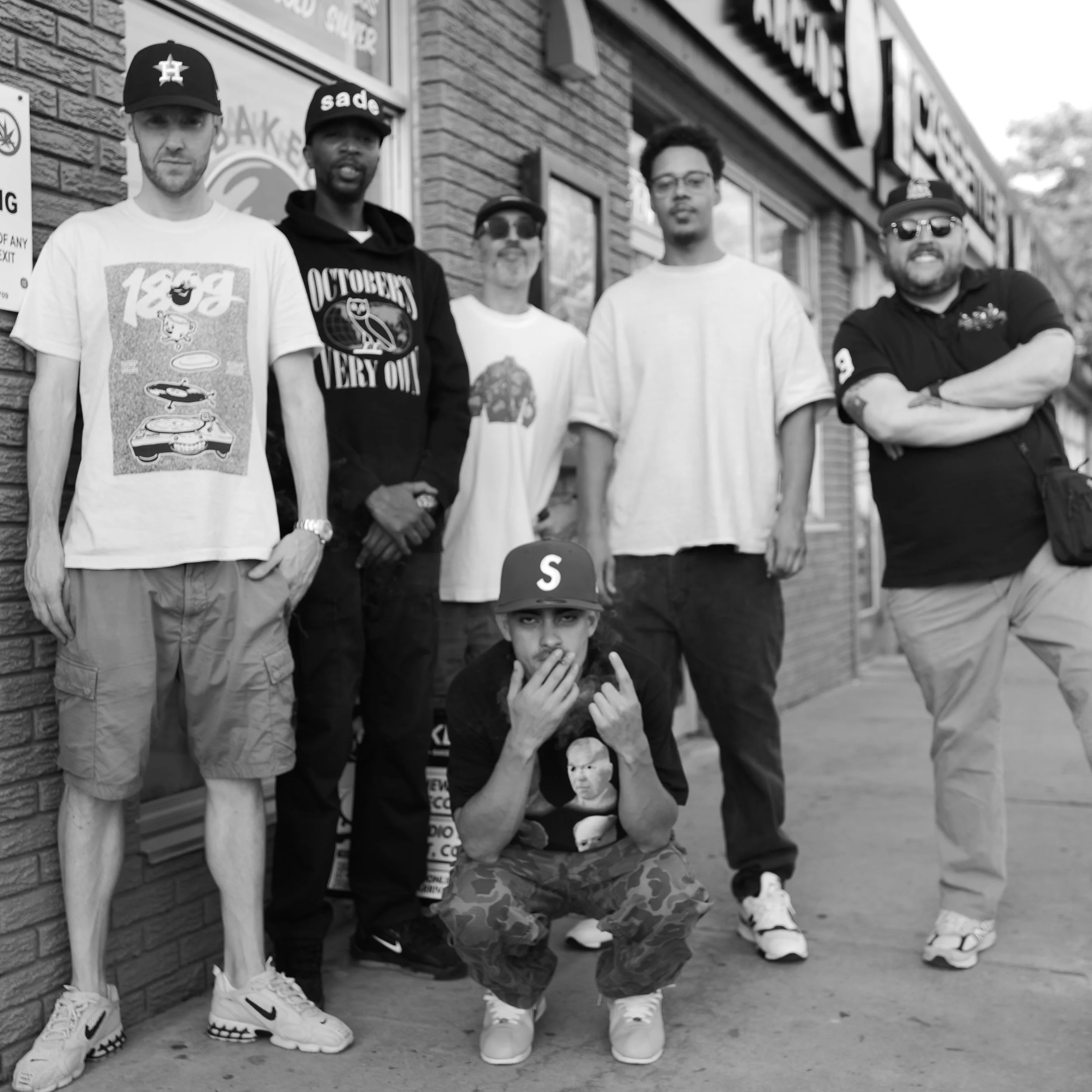 Six men pose on a city sidewalk in front of a brick building.