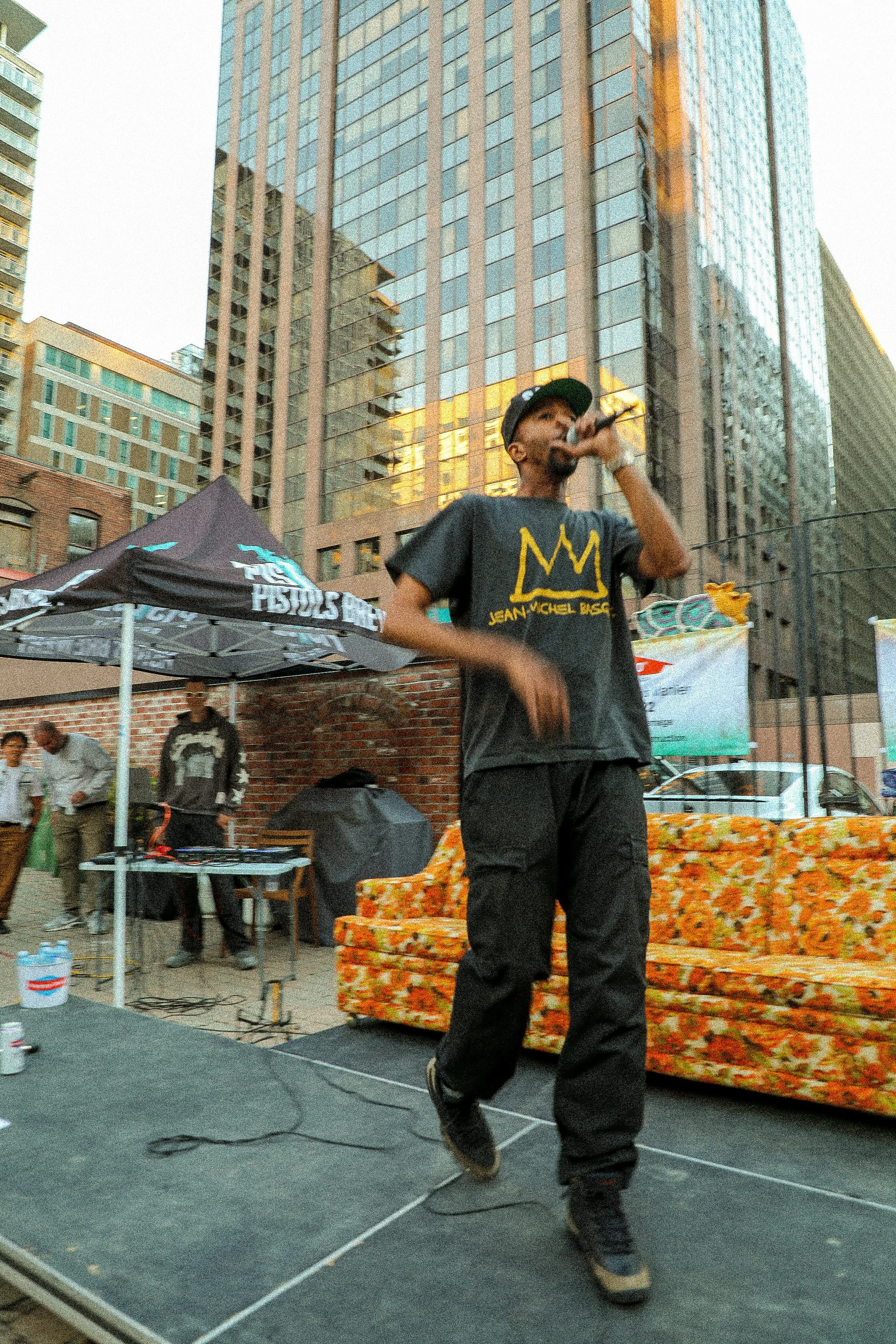 A man wearing a black t-shirt with a yellow crown graphic and black pants is performing on an outdoor stage in a city, holding a microphone. Tall buildings and an audience are visible in the background.