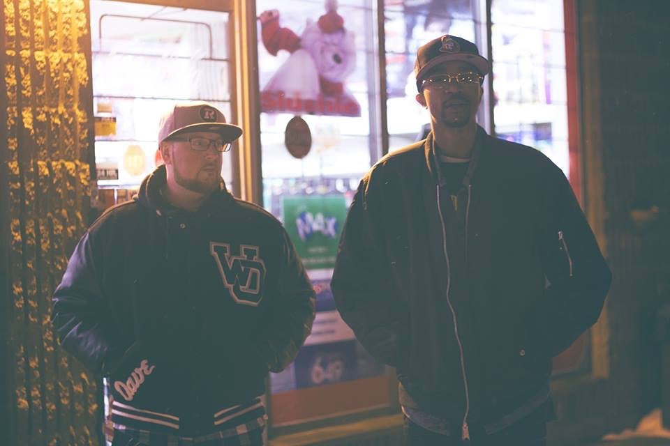 Two men wearing black jackets and baseball caps standing outside a window at night, with neon signs and TV screens in the background.
