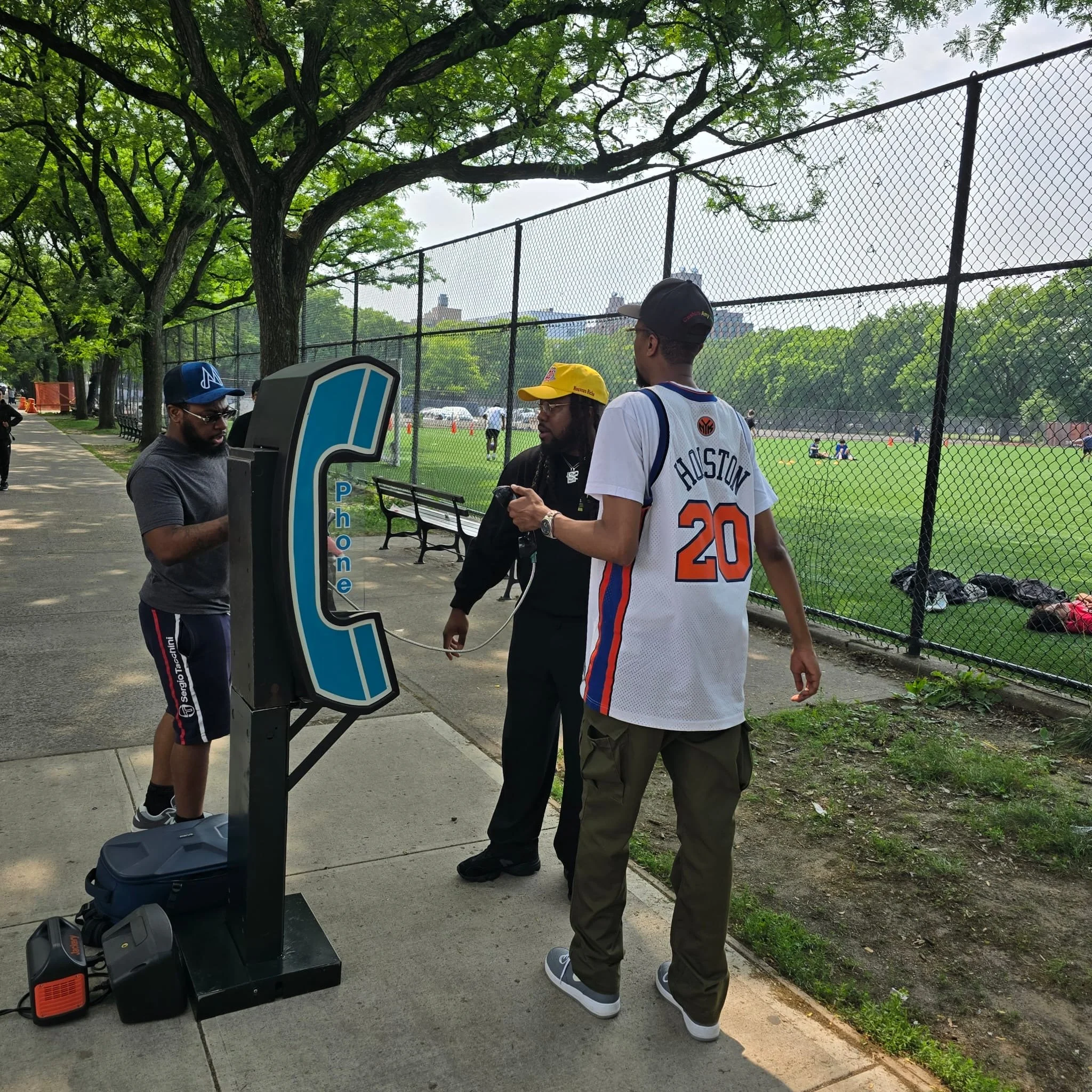 Three men talking on a sidewalk near a pay phone booth, with a park and a baseball field behind a chain-link fence in the background.