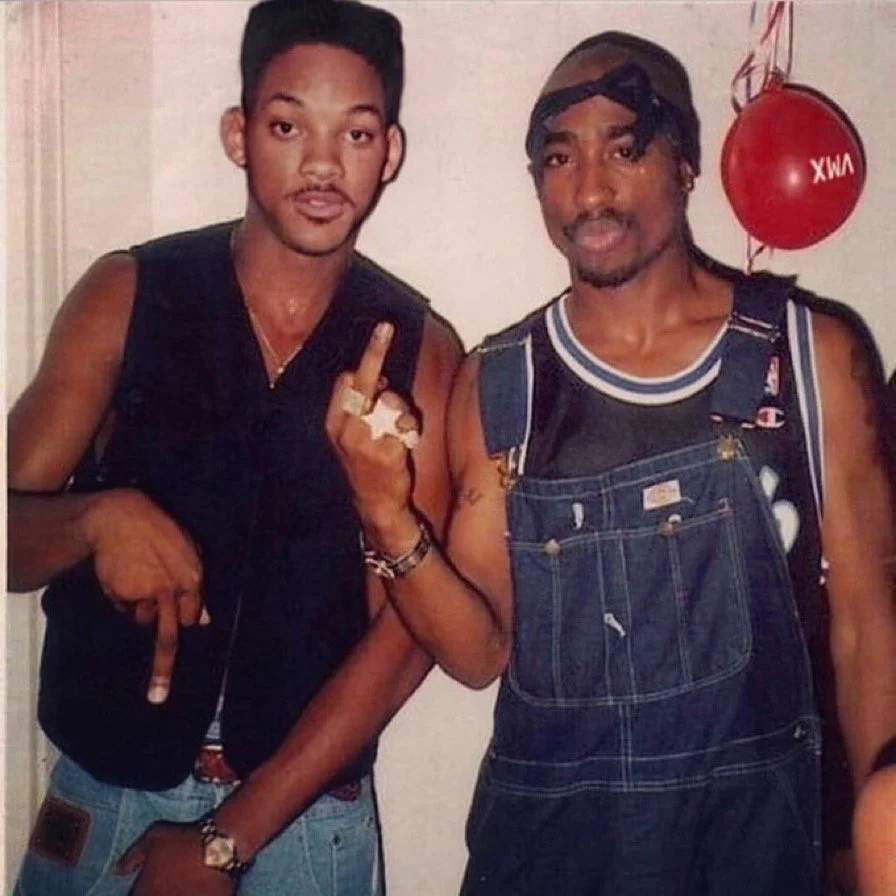 Two men standing together, one showing his middle finger and the other with a serious expression, in casual clothing, with a red balloon in the background.