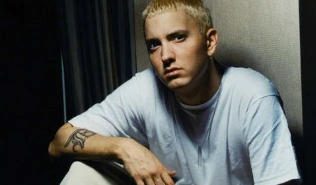 Young man with blond hair and tattoos on his arm, wearing a white shirt, sitting indoors against a dark background.