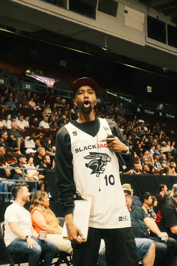 A man in a basketball jersey and cap stands in a crowded sports arena, holding a clipboard and pointing to himself.