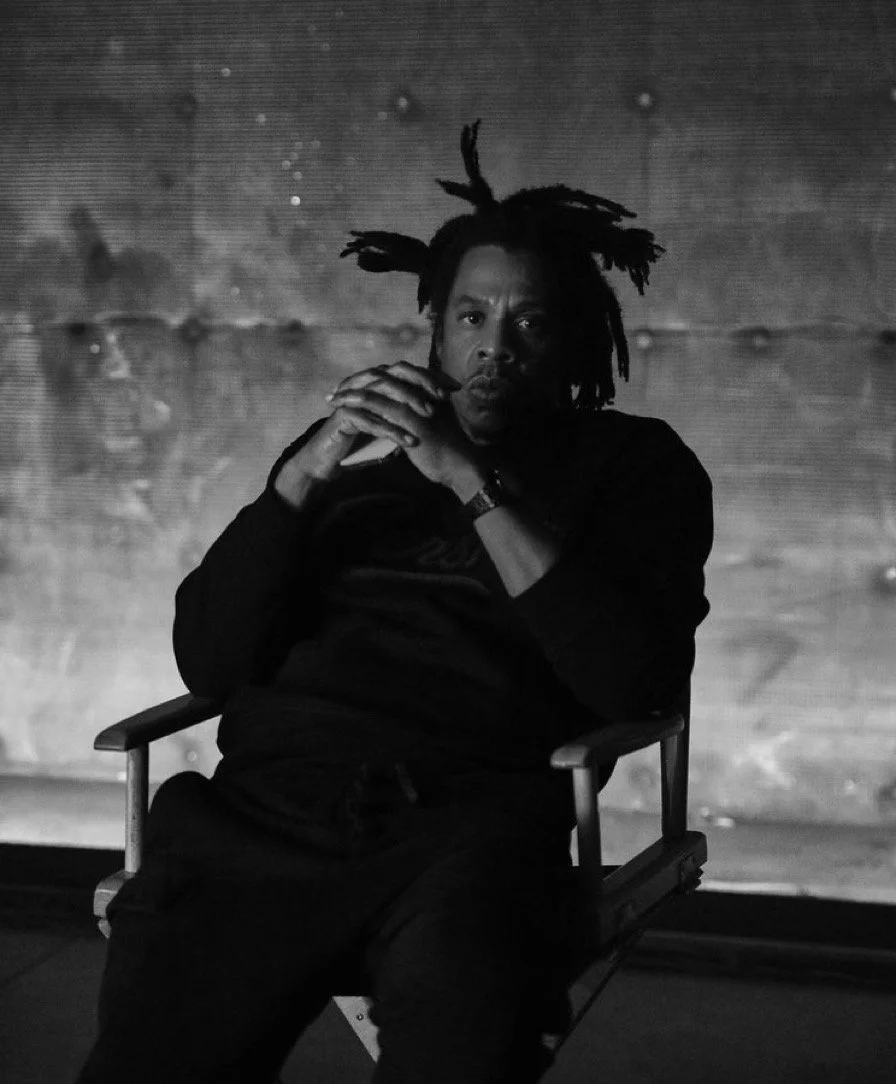 Black and white photo of a man with dreadlocks sitting on a chair, looking at the camera, with his hands clasped together. Background appears to be textured and illuminated.