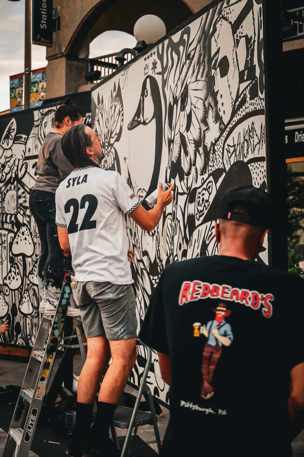 Two artists work on a large black and white mural with floral and animal designs on a city street, while a person in the foreground wears a black t-shirt with a 'Redbeard's' logo and a character holding a beer.