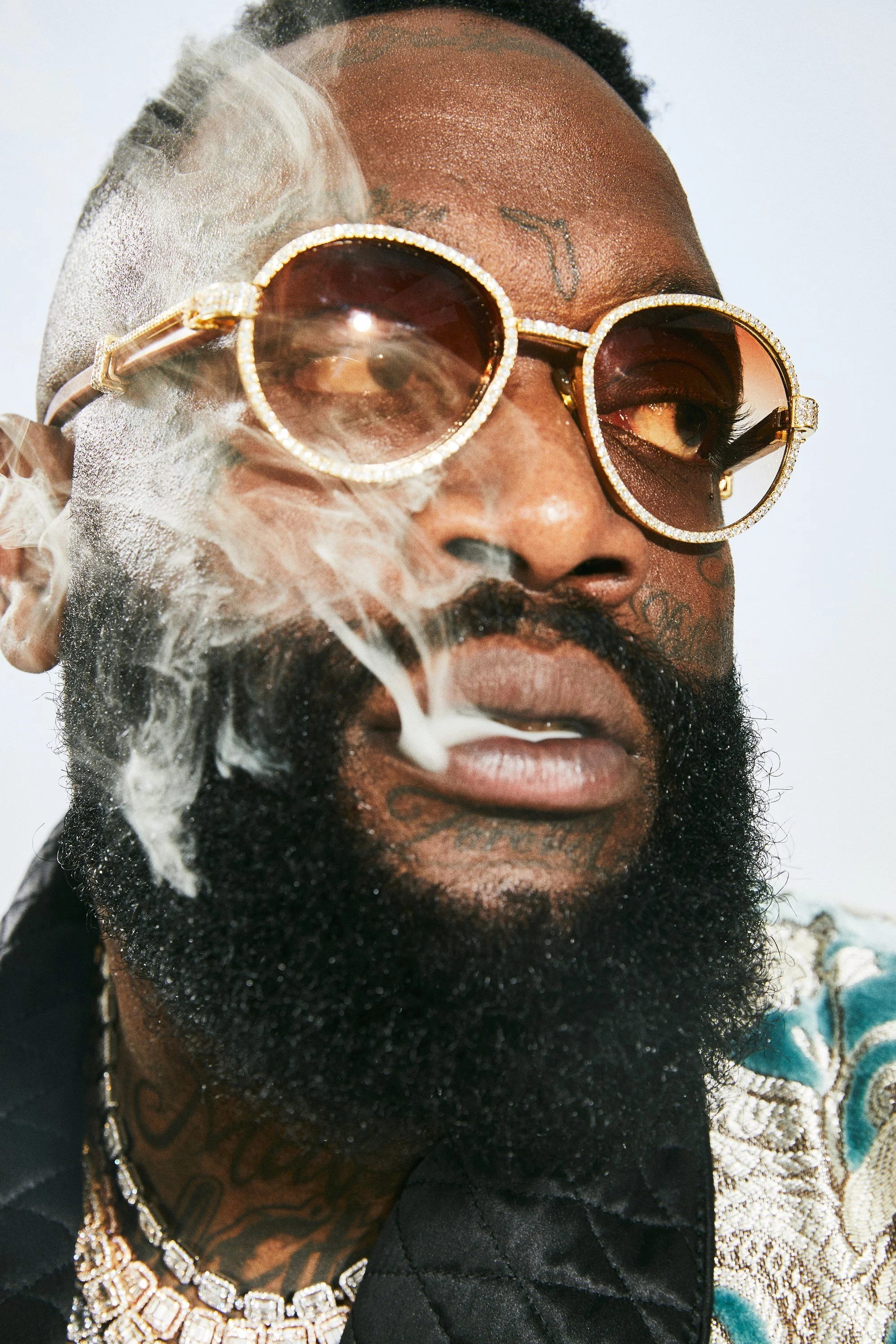 Close-up of a man with sunglasses, tattoos, a full beard, and smoking, with smoke around his face and glowing highlights.