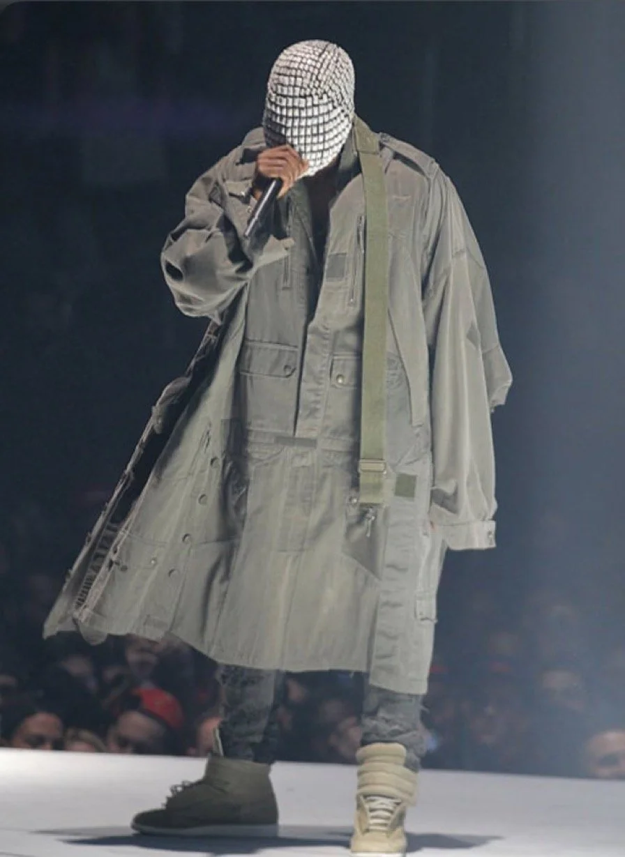 A person on a runway wearing a large gray coat, camouflage pants, beige footwear, and a patterned cap, holding a microphone and bowing their head.