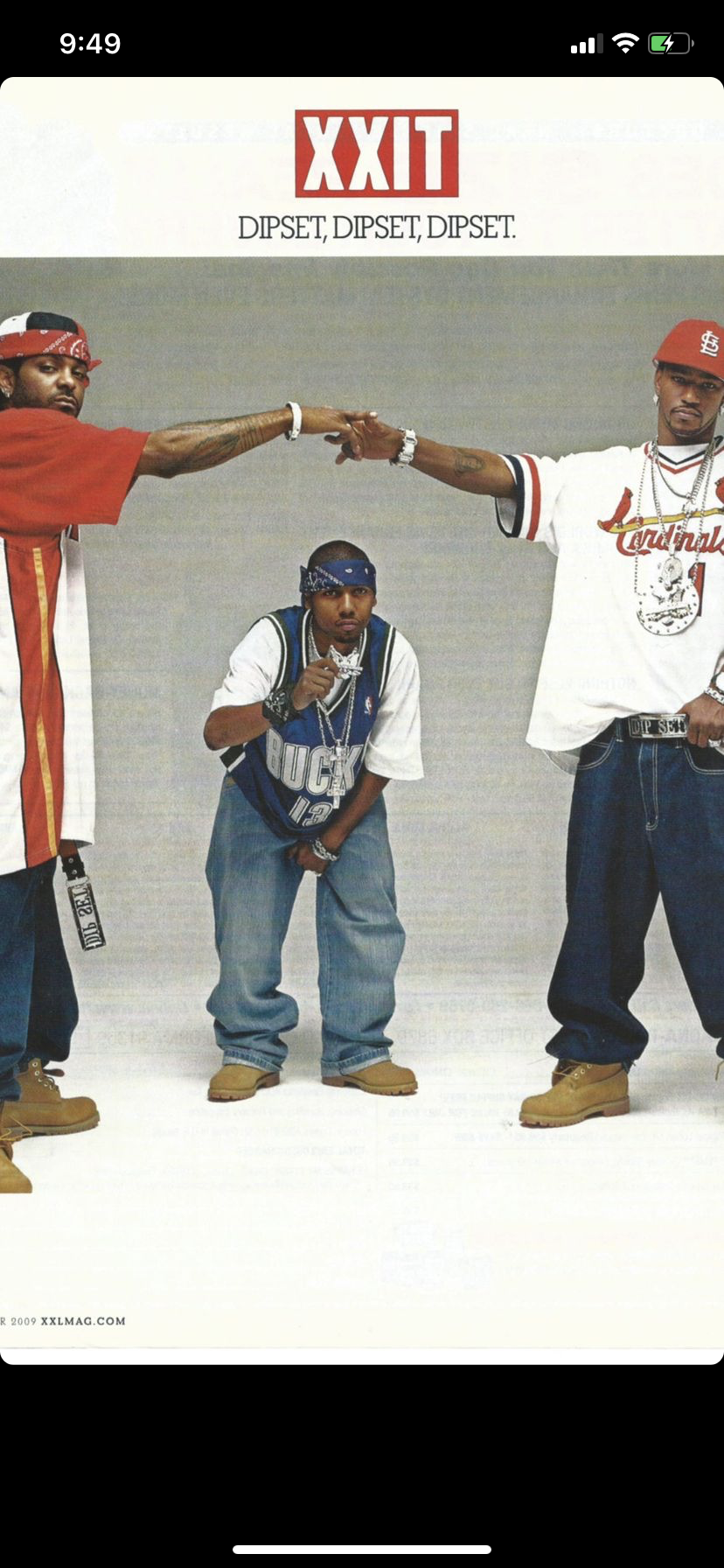 Three young men posing together, two of them pointing towards each other with their fingers in a cross, with a white background. One man is in the middle, kneeling and holding a chain to his mouth. The other two are standing, wearing casual streetwea