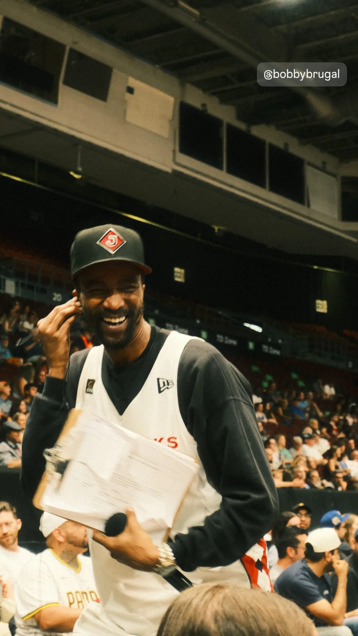 A man smiling, holding a microphone and some papers, wearing a black and white sports jersey and a black cap with a red logo, at an indoor sports event with an audience in the background.