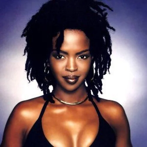 Portrait of a woman with dark skin, curly dreadlock hair, wearing a black top and gold jewelry against a dark background.