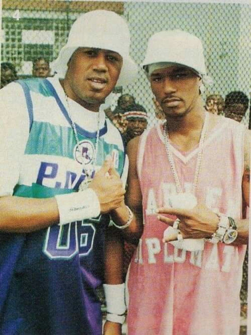 Two men dressed in basketball jerseys with headbands and jewelry, making hand gestures, standing among a group of people.