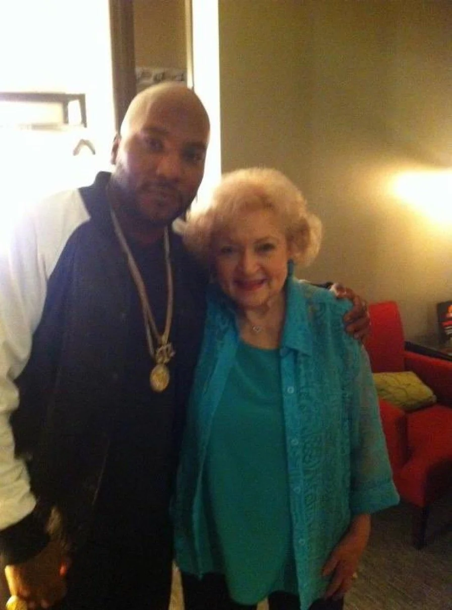 A man and an elderly woman standing close together, with the man wearing a gold chain and a black and white jacket, and the woman wearing a blue shirt with an embroidered blue jacket, in a dimly lit room with red chairs in the background.