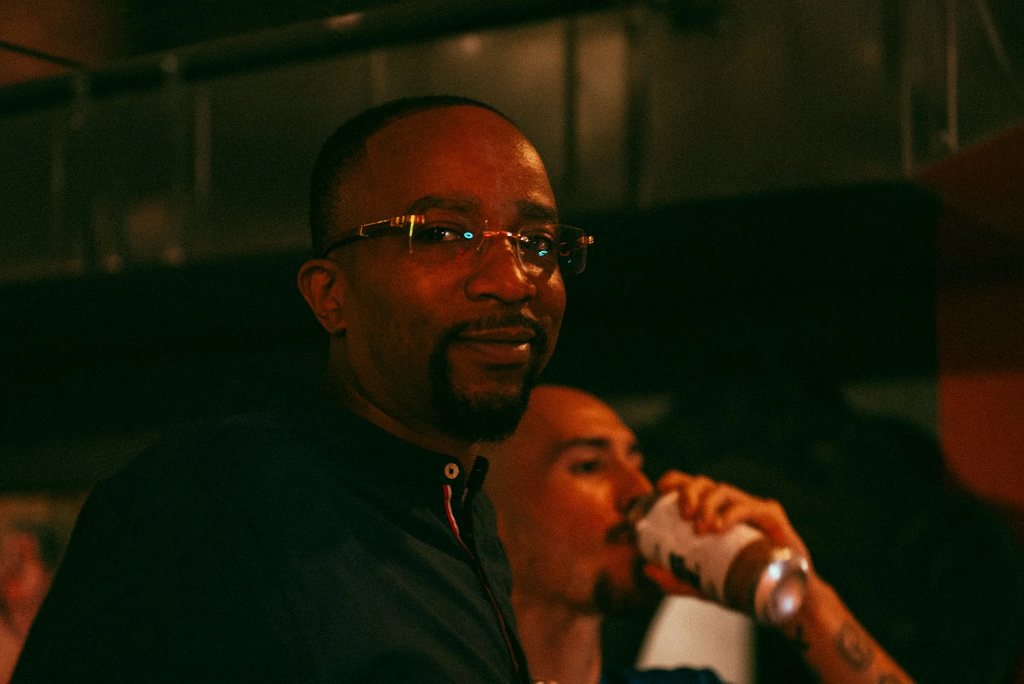 A man with glasses and a goatee looking at the camera, in a dimly lit setting, with another man drinking from a can in the background.