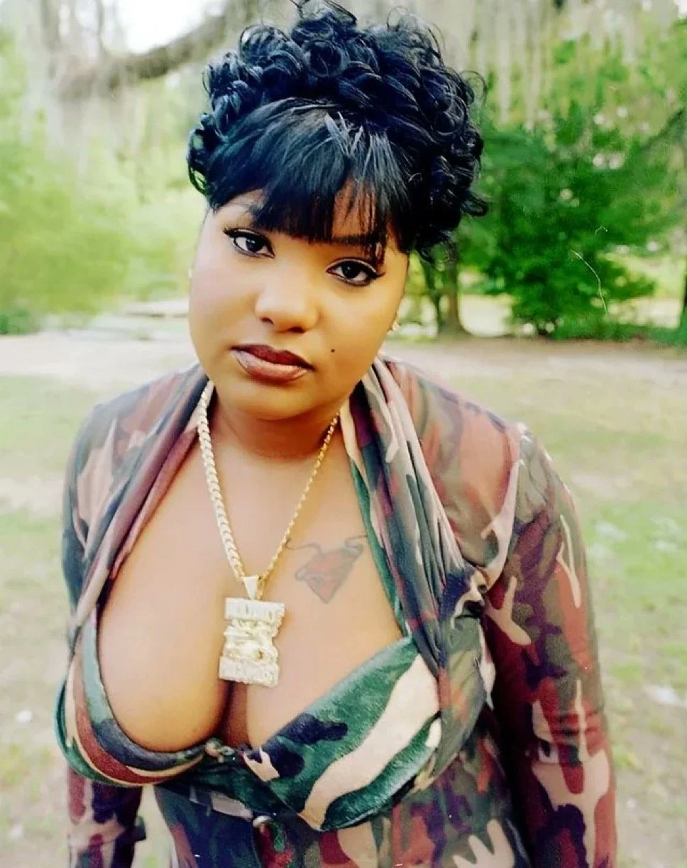 Close-up of a woman with short curly black hair, wearing a camouflage jacket, gold chain with a large pendant, and a green patterned dress, standing outdoors with trees and greenery in the background.