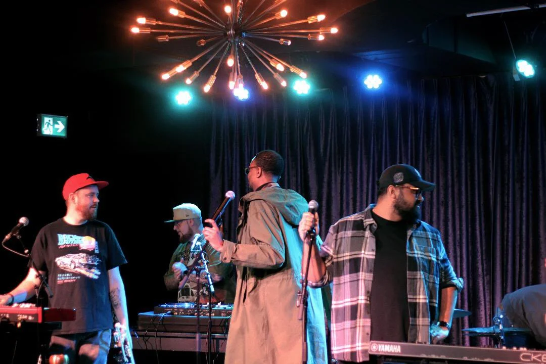 Four men on stage with microphones, a DJ, and musical equipment under colorful stage lighting and a dark curtain backdrop.