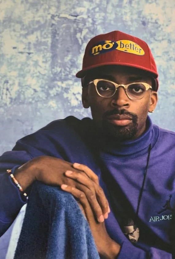 Person wearing a red cap with yellow text, glasses, a purple Nike sweatshirt, and sitting against a mottled blue background.