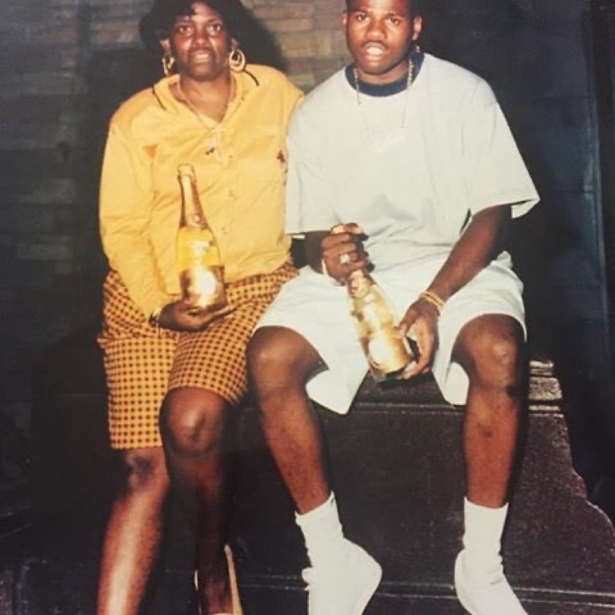 A woman and a young man sitting together holding beer bottles, with the woman wearing a yellow top and checkered skirt, and the man dressed in a gray t-shirt and white shorts.