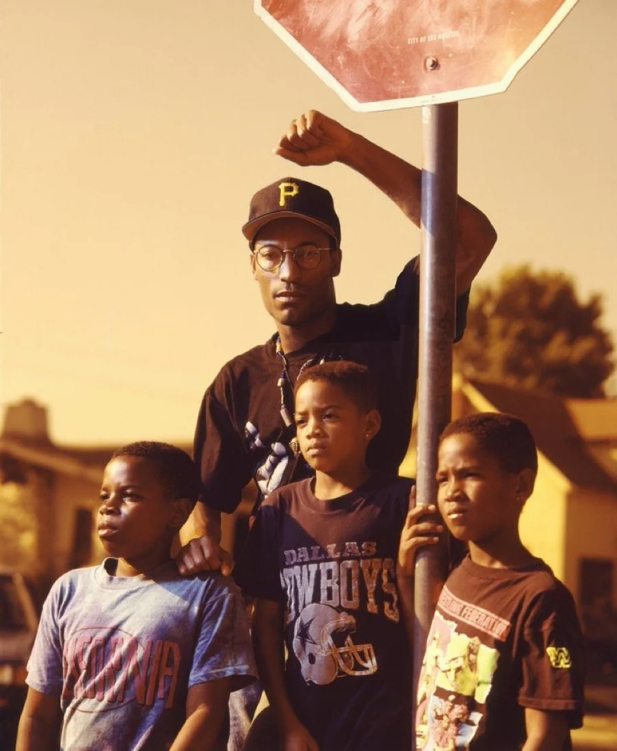 A young man wearing a black t-shirt, glasses, and a black cap with a yellow letter 'P' stands behind three boys, under a stop sign, on a neighborhood street during sunset. The boys wear casual t-shirts, and the young man has a serious expression.