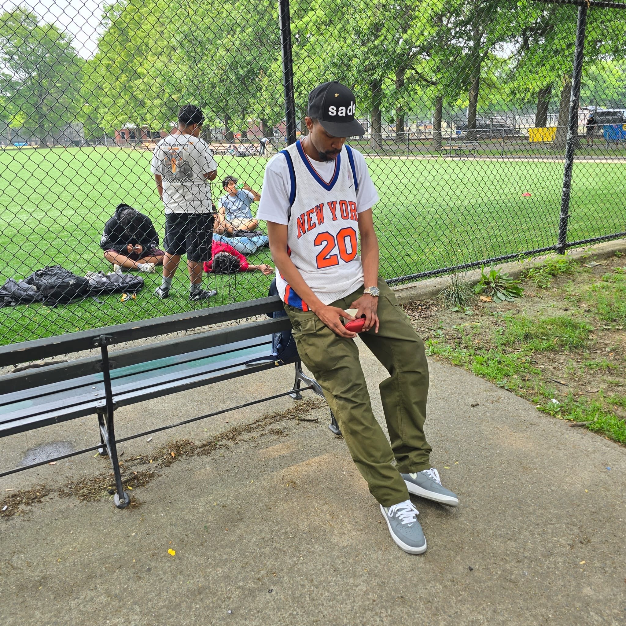 A man wearing a black cap, white Knicks basketball jersey with the number 20, khaki pants, and gray sneakers is sitting on a park bench, looking down at his red phone. In the background, across a fence, some people are sitting on the grass, and other