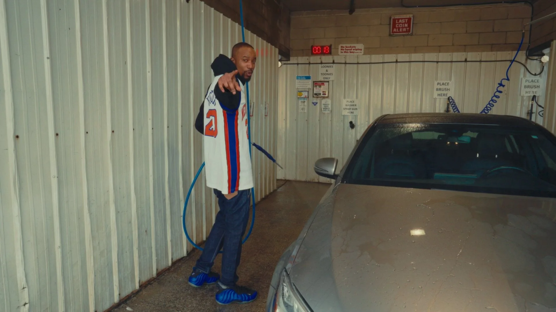 A man standing inside a car wash bay pointing at the camera while washing a silver car with a hose.