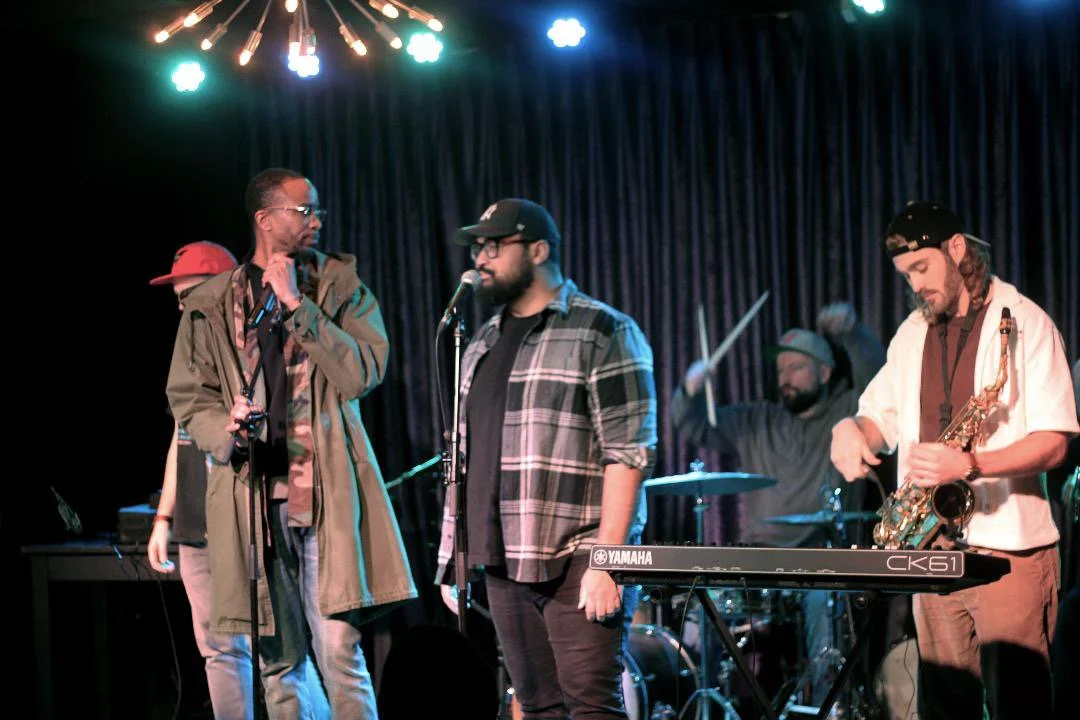 A group of five male musicians preparing to perform on a stage with a keyboard and drum set, one man is holding a microphone, others are holding instruments, with stage lights overhead and a dark curtain background.