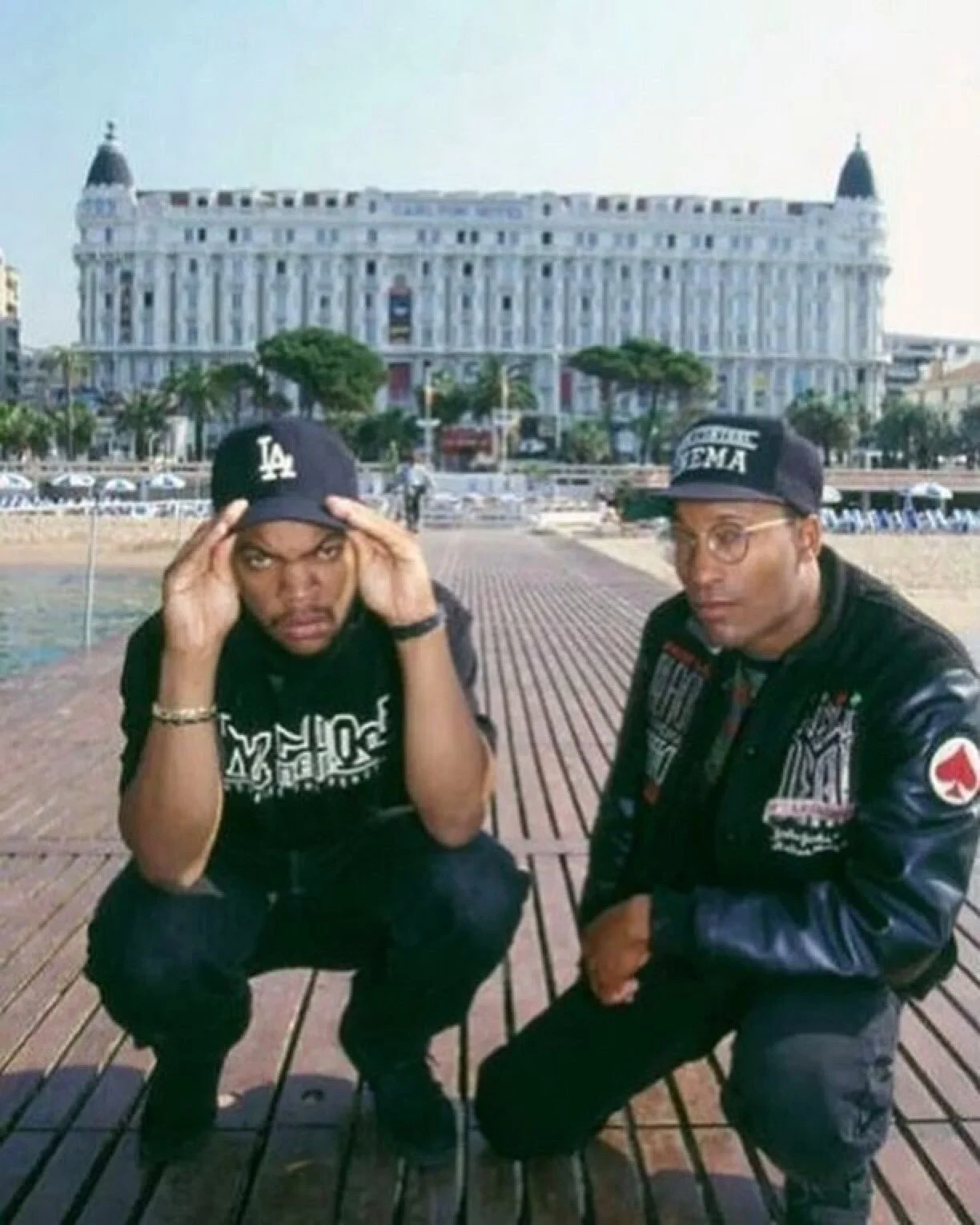 Two men squatting on a wooden boardwalk by the beach, with a large ornate white building in the background. Both men wear black caps and casual clothing.