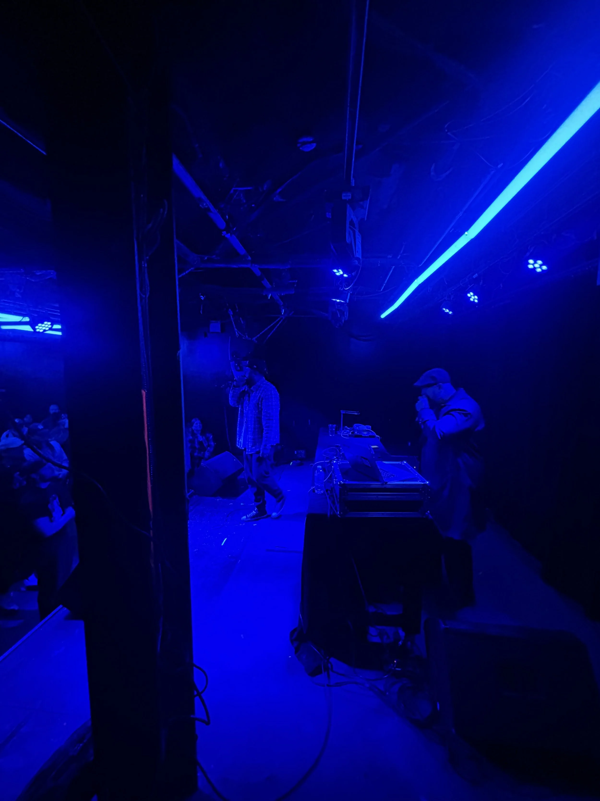 A dimly lit stage with blue lighting, featuring a person with a microphone, a DJ or music setup, and several audience members watching.